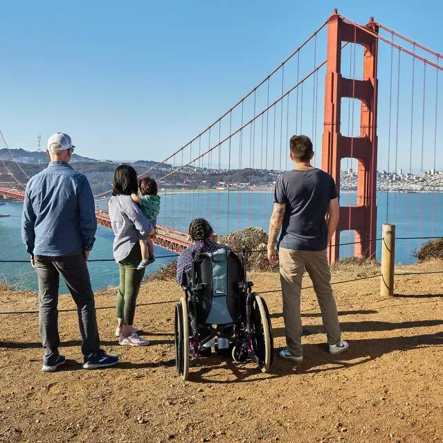 휠체어를 탄 사람 한 명을 포함한 사람들의 그룹이 마린 헤드랜드에서 Golden Gate Bridge 바라보고 있는 모습이 뒤쪽에서 보입니다.