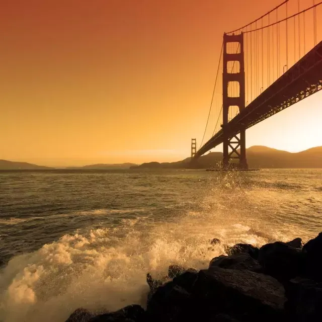 Waves crash near Fort Point beneath the Golden Gate Bridge at sunset.